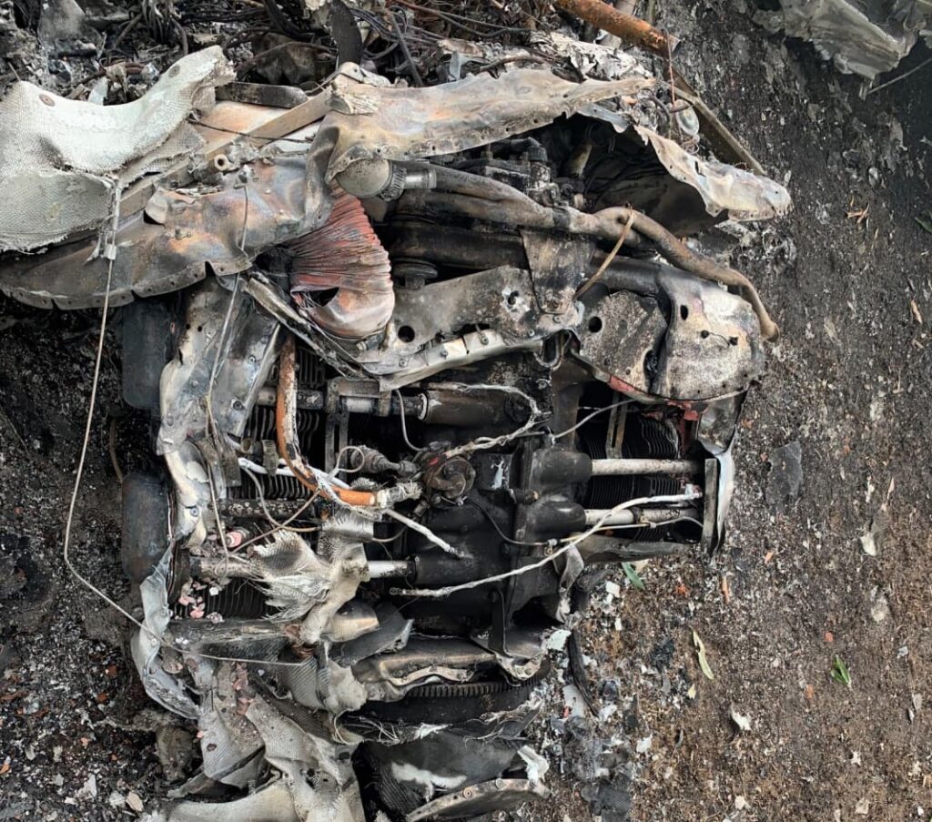 Close-up of the burned engine of the aircraft, showing melted and deformed metal, exposed cylinders, and fire-damaged wiring. The engine is embedded in darkened soil with ash and debris scattered around it.