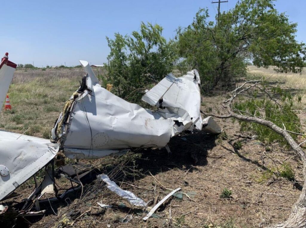 Damaged section of the Cessna’s fuselage lying on the ground. The tail section is bent and twisted with torn metal and wiring visible, and nearby trees and brush are broken from the impact.