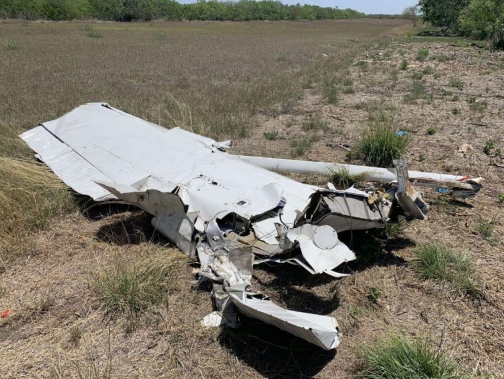 Detached wing from the Cessna lying in a field. The metal is heavily crumpled with pieces of the control surfaces damaged, and the surrounding area is dry grass and scattered debris.