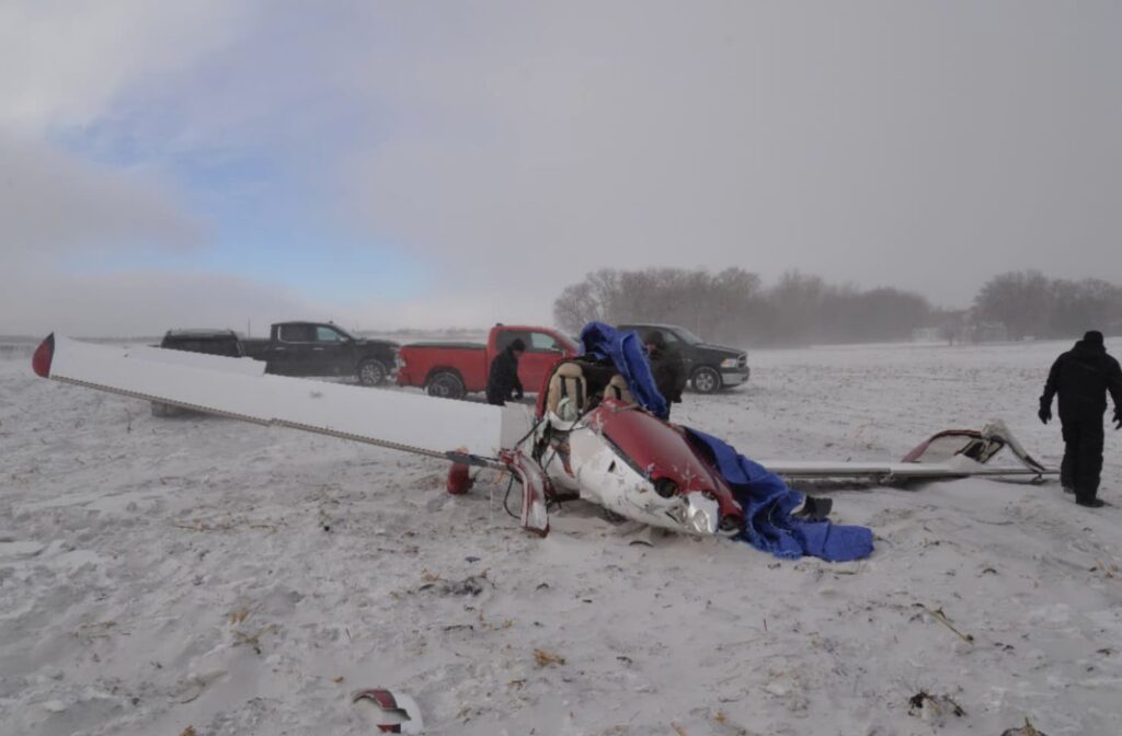 The wreckage of the same Cirrus SR22 rests in a snow-covered field. The tail section is broken, and a tarp partially covers the cabin. Several pickup trucks and people in winter clothing stand nearby under low gray clouds and blowing snow.