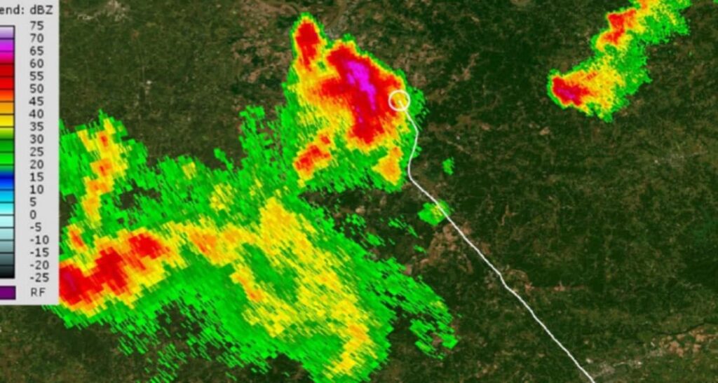 Weather radar map showing a white flight track line leading into an area of intense precipitation. The strongest storm cell is displayed in red and purple near the track’s endpoint, with yellow and green areas surrounding it. A color scale on the left shows dBZ intensity levels.
