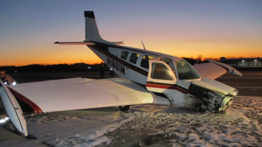 A Beechcraft A36 Bonanza, registration N1861M, resting on its nose at sunset on a runway. The aircraft shows significant damage to the nose and propeller area, with firefighting foam visible on the pavement around it.