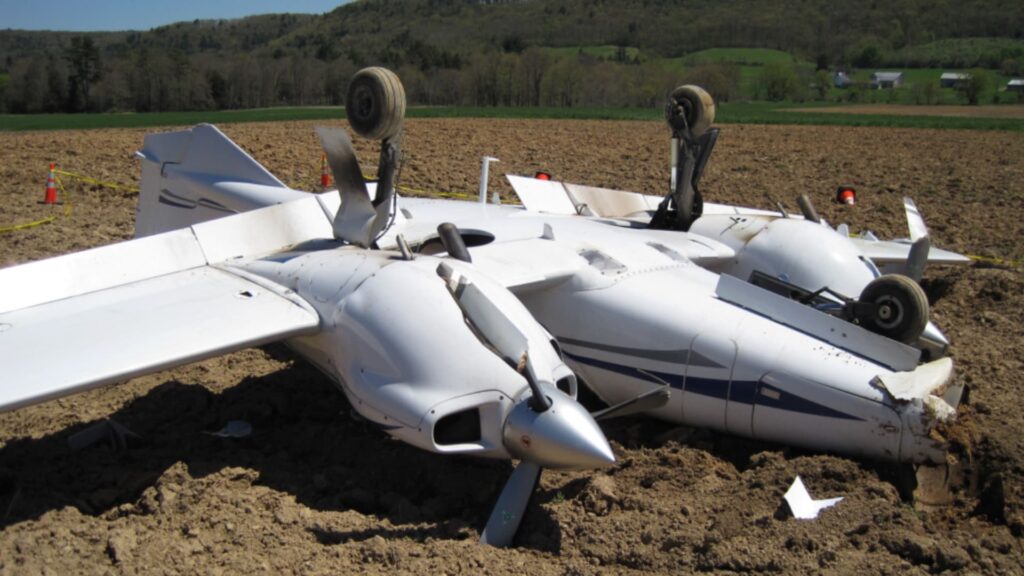 A Diamond DA42 twin-engine airplane overturned in a plowed field after a forced landing, showing significant damage with the landing gear extended upward and both propellers bent.