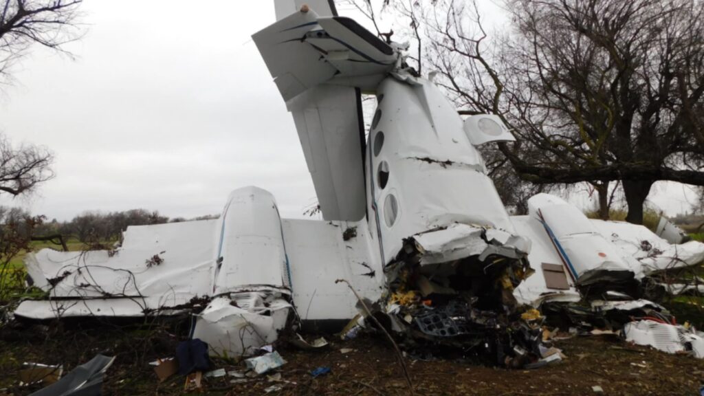 Rear view of Cessna 414 N4765G wreckage showing crushed fuselage and engines, with tail section upright and wings torn, surrounded by broken parts and debris in a wooded field.