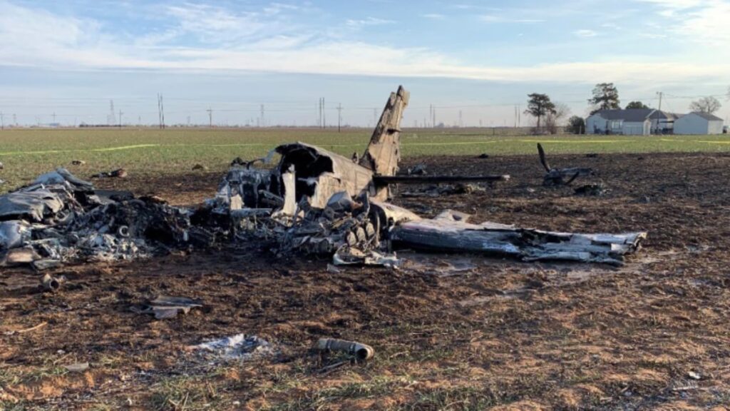Burned wreckage of a small twin-engine airplane resting in an open field. The fuselage and wings are heavily damaged and partially consumed by fire, with debris scattered around the impact area under a clear sky.