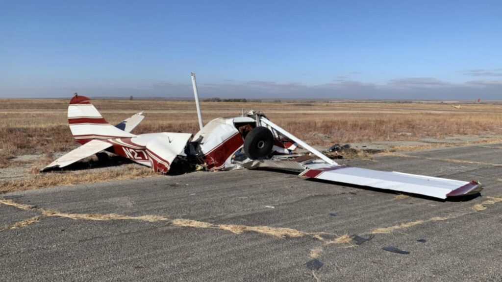 Side view of the Cessna 170B N2710D wreckage on an airport taxiway. The fuselage and wings are severely buckled, the nose crushed, and one main landing gear still attached while the propeller lies broken nearby.