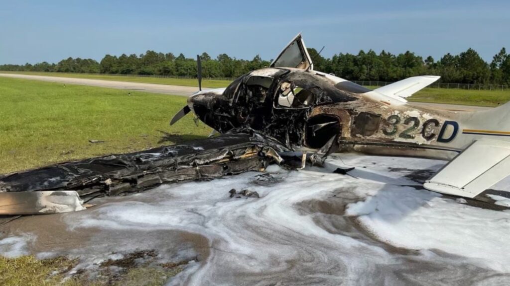 Burned Cirrus SR22 aircraft resting beside a runway after a fire, with the fuselage and left wing severely damaged and covered in firefighting foam. The registration number “N432CD” is partially visible on the tail, and the aircraft’s interior and nose section are completely destroyed. The photo shows clear skies, green grass, and the paved runway in the background.