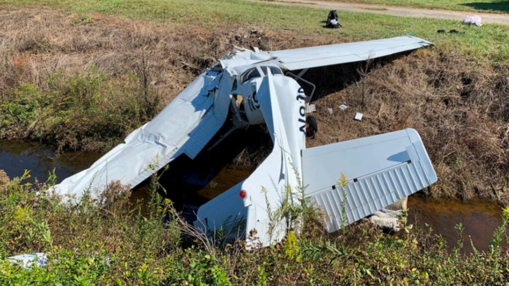 Damaged Cessna 172P aircraft lying in a ditch beside a grassy area with two vehicles parked nearby. The wings are bent and twisted, and debris from the crash is scattered around the site.