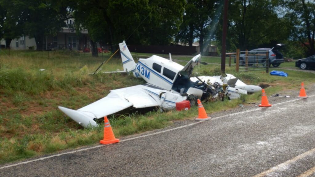 A wrecked white twin-engine airplane, N473M, lies on the edge of a country road with major damage to the nose and wings. Orange cones mark the scene, and a few vehicles and a farmhouse are visible in the background among trees and grass.