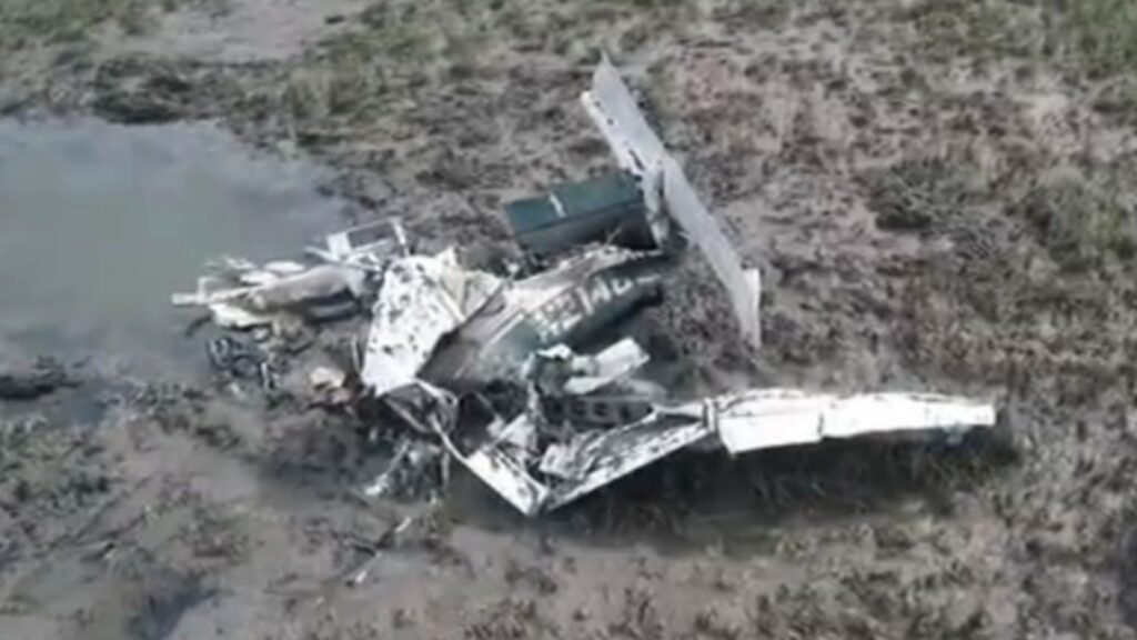 Aerial view of a small airplane wreckage lying in a muddy marsh beside a shallow pool of water. The fuselage and wings are heavily damaged and fragmented, with debris scattered across the wet ground.
