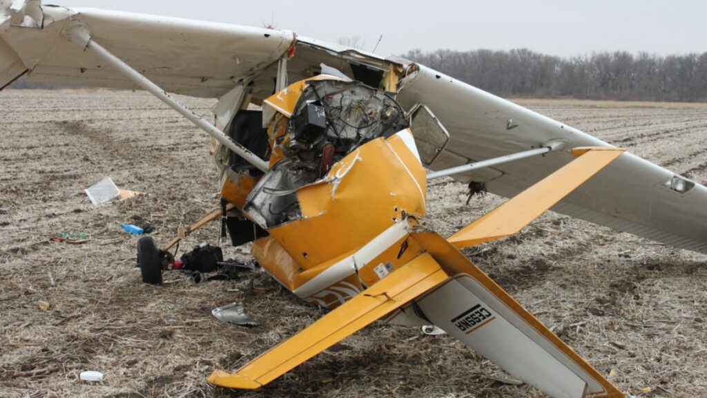 Right-side view of a yellow and white Cessna 150 crash site in a farm field. The nose and cockpit area are heavily damaged, with the “Cessna” logo visible on the tail and the wings bent from impact.