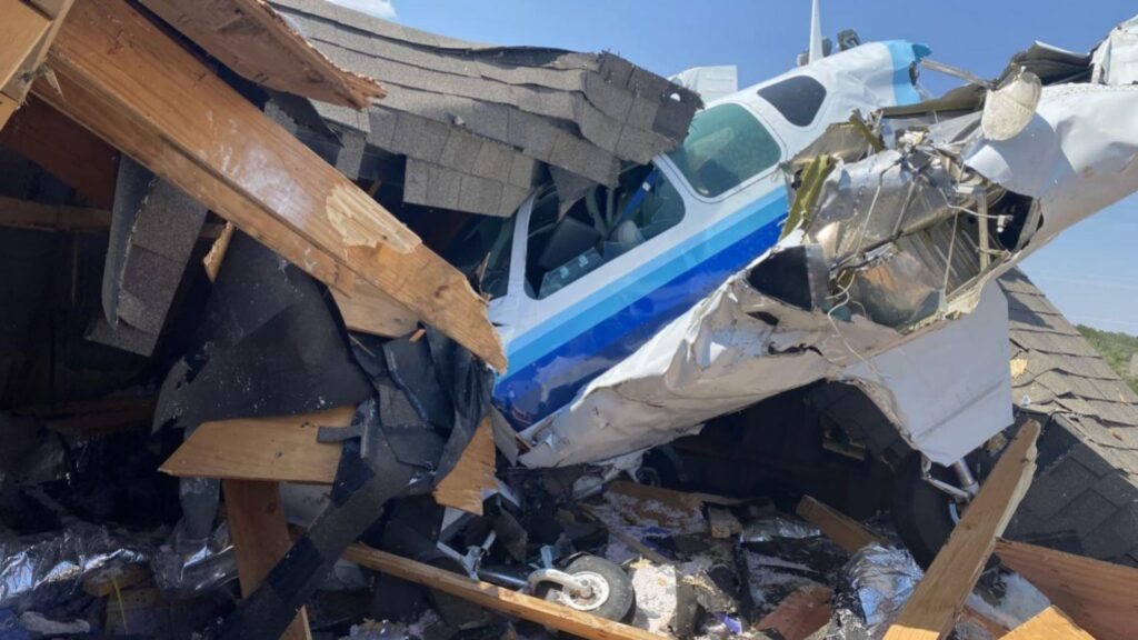 Close-up of a damaged Beechcraft Bonanza aircraft embedded in the roof of a two-story house, with splintered wooden beams and roofing debris surrounding the fuselage under clear blue skies.