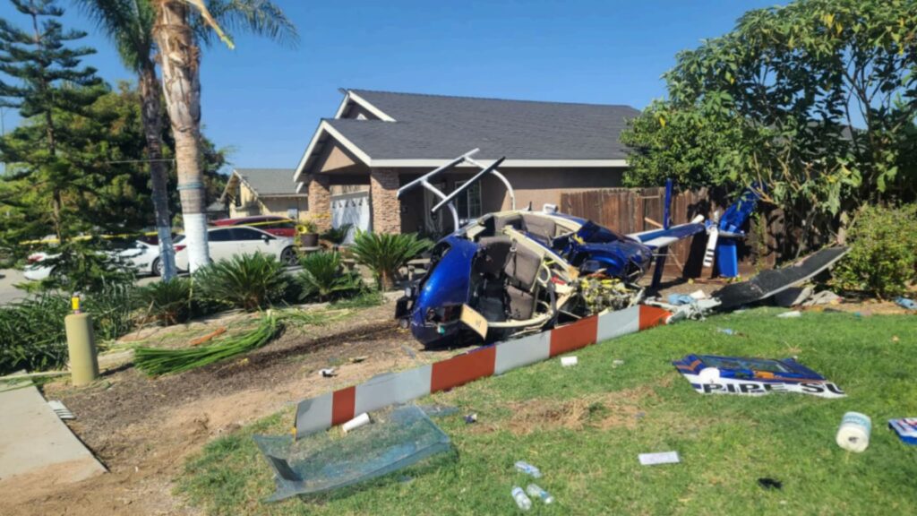 A blue Bell 206B helicopter lies heavily damaged in the front yard of a suburban home. The fuselage is broken open, the tail section is twisted and separated, and debris is scattered across the grass and driveway. A house, trees, and parked cars are visible in the background under clear skies, with police tape marking off the area.