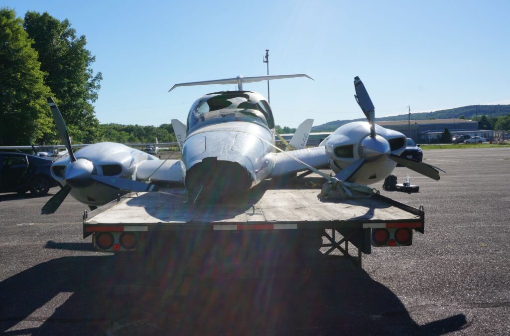 The wreckage of a Diamond DA42 loaded on a flatbed trailer, showing a crushed nose section and bent propellers, with the wings and fuselage visibly damaged.
