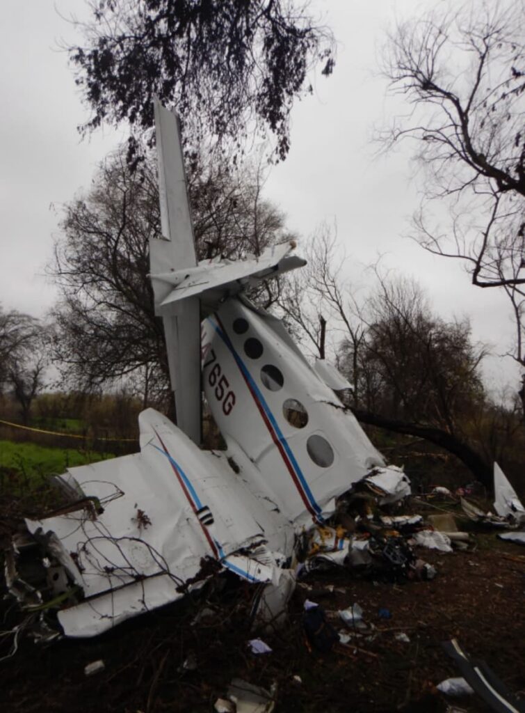 Wreckage of Cessna 414 N4765G standing nearly vertical against trees, with the tail section intact but fuselage crushed at the base, surrounded by debris in a wooded area.