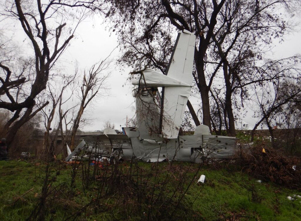 Side view of Cessna 414 N4765G wreckage with tail section upright among leafless trees, fuselage heavily damaged and broken, with debris scattered across grass near a roadway.