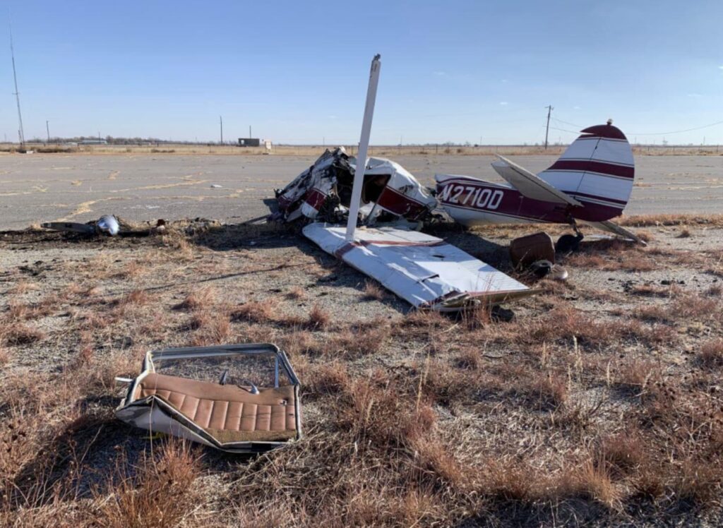 Wreckage of a Cessna 170B N2710D lying inverted on the edge of an airport ramp. The fuselage is crushed and broken forward of the cabin, with the right wing detached and a seat lying in the foreground amid dry grass.