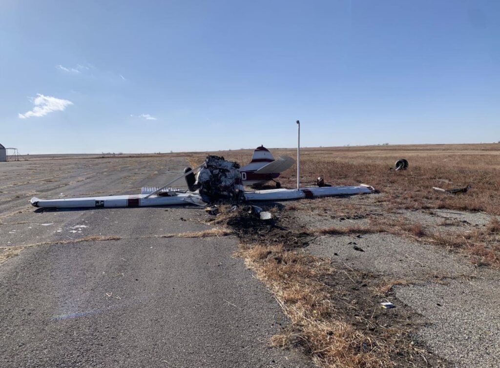 Front view of the Cessna 170B N2710D crash site showing the destroyed engine area, separated propeller, and debris trail leading onto a ramp surrounded by open prairie terrain under clear skies.