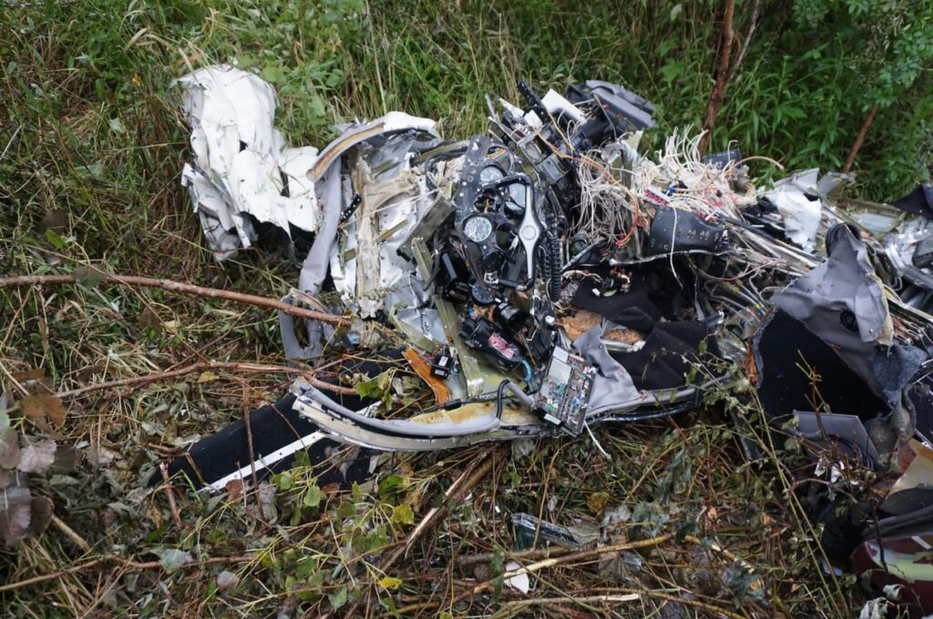 Close-up view of the destroyed cockpit and instrument panel of the aircraft. Wires, gauges, and avionics components are tangled and torn apart, surrounded by vegetation. The wreckage shows severe crushing and fragmentation from the impact.