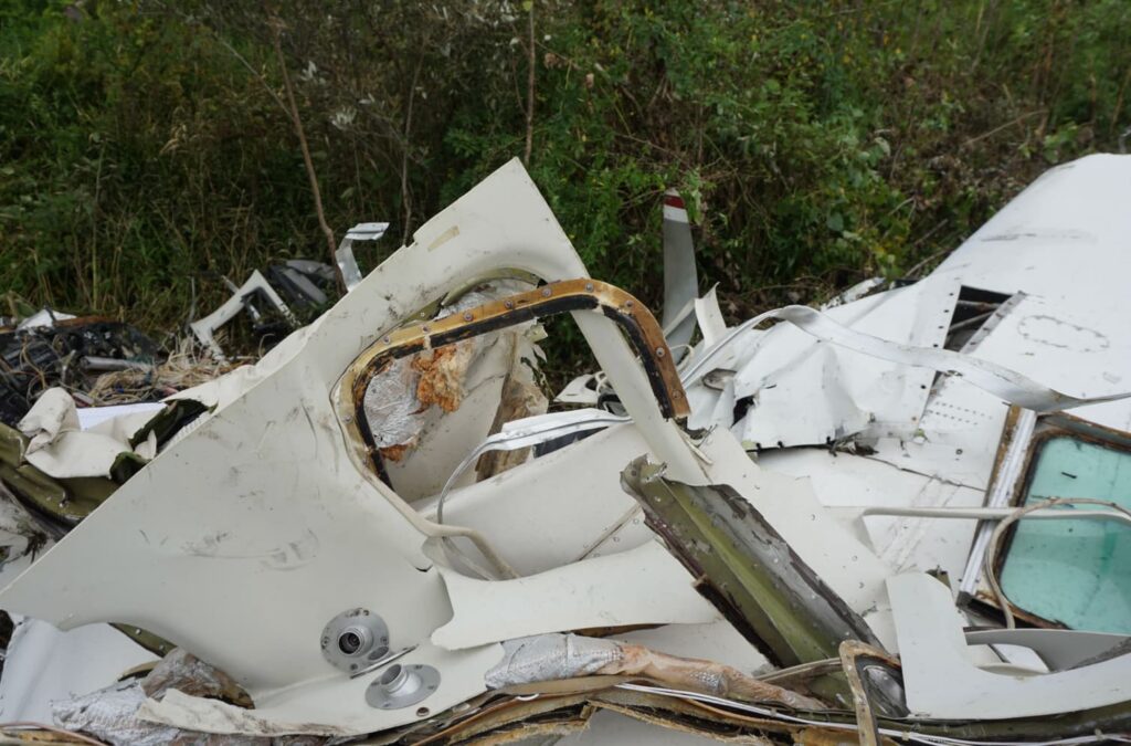 A detailed photo of a torn aircraft fuselage section, showing the damaged cabin door frame and interior insulation. The metal skin is twisted and peeled back, exposing structural layers and wiring, with nearby brush and vegetation encroaching on the debris.