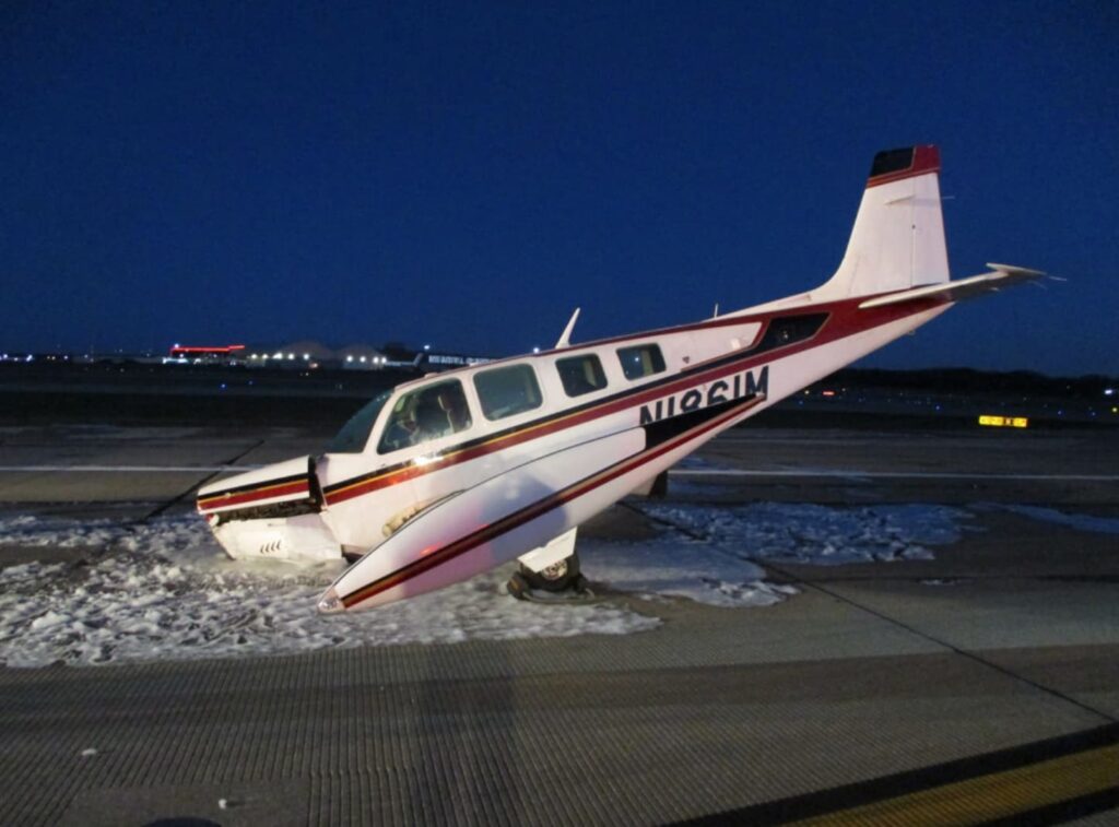 Side view of a damaged Beechcraft A36 Bonanza, registration N1861M, at night on an airport runway. The airplane’s nose is crushed, the propeller bent, and foam surrounds the area where the aircraft came to rest.