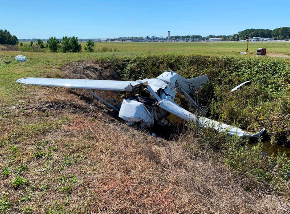 Cessna 172P crash site near Newport News airport showing the aircraft crumpled in a drainage ditch. The nose and cabin area are crushed, the wings are deformed, and the airport control tower is visible in the distance.