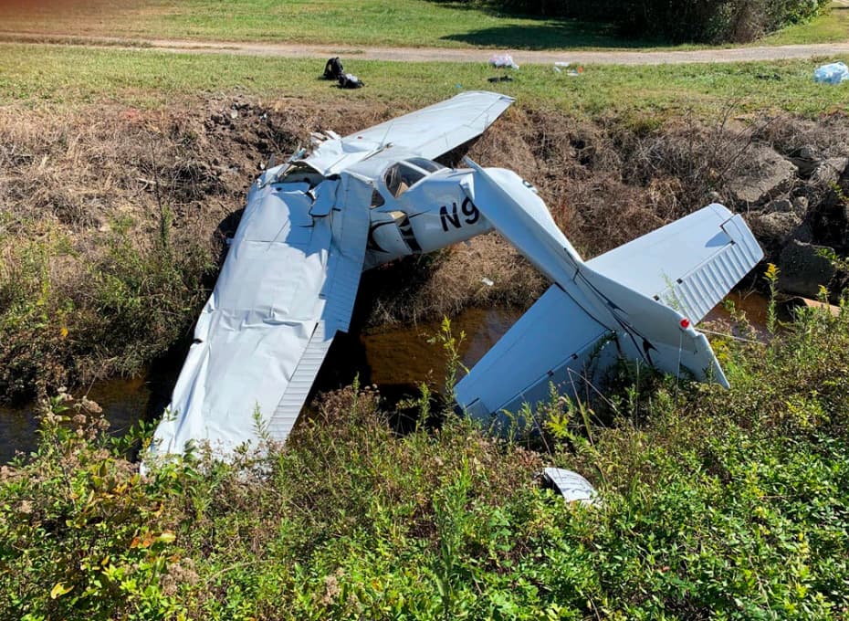 Cessna 172P wreckage resting in a shallow drainage ditch with severe damage to both wings and fuselage. The aircraft’s tail is elevated while the fuselage is partially submerged, and grass surrounds the ditch.