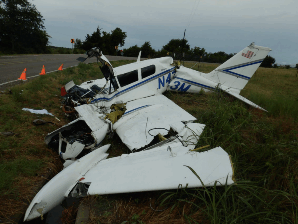 A wrecked white twin-engine airplane, N473M, lies on the edge of a country road with major damage to the nose and wings. Orange cones mark the scene, and a few vehicles and a farmhouse are visible in the background among trees and grass.