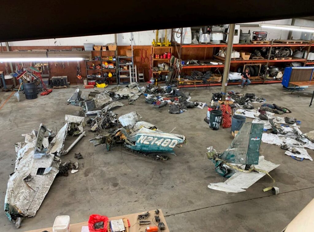 Wreckage of a Piper PA-28 aircraft labeled “N21480” laid out inside a hangar for examination. Pieces of the fuselage, wings, and tail are arranged on the floor amid tools, bins, and maintenance equipment.