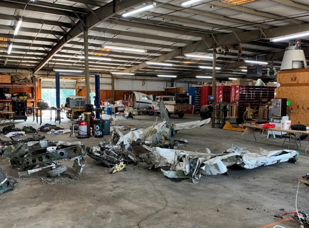 Interior view of an aircraft maintenance hangar with the disassembled remains of a Piper PA-28 aircraft spread across the floor. The damaged airframe parts are organized for inspection among workbenches and storage shelves.
