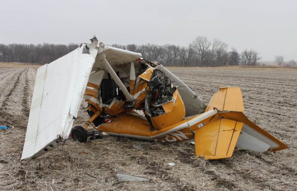 Wreckage of a yellow and white Cessna 150 lying in a harvested field near Auburn, Nebraska. The fuselage is severely crushed, and the left wing is folded over the cabin, showing the impact damage.
