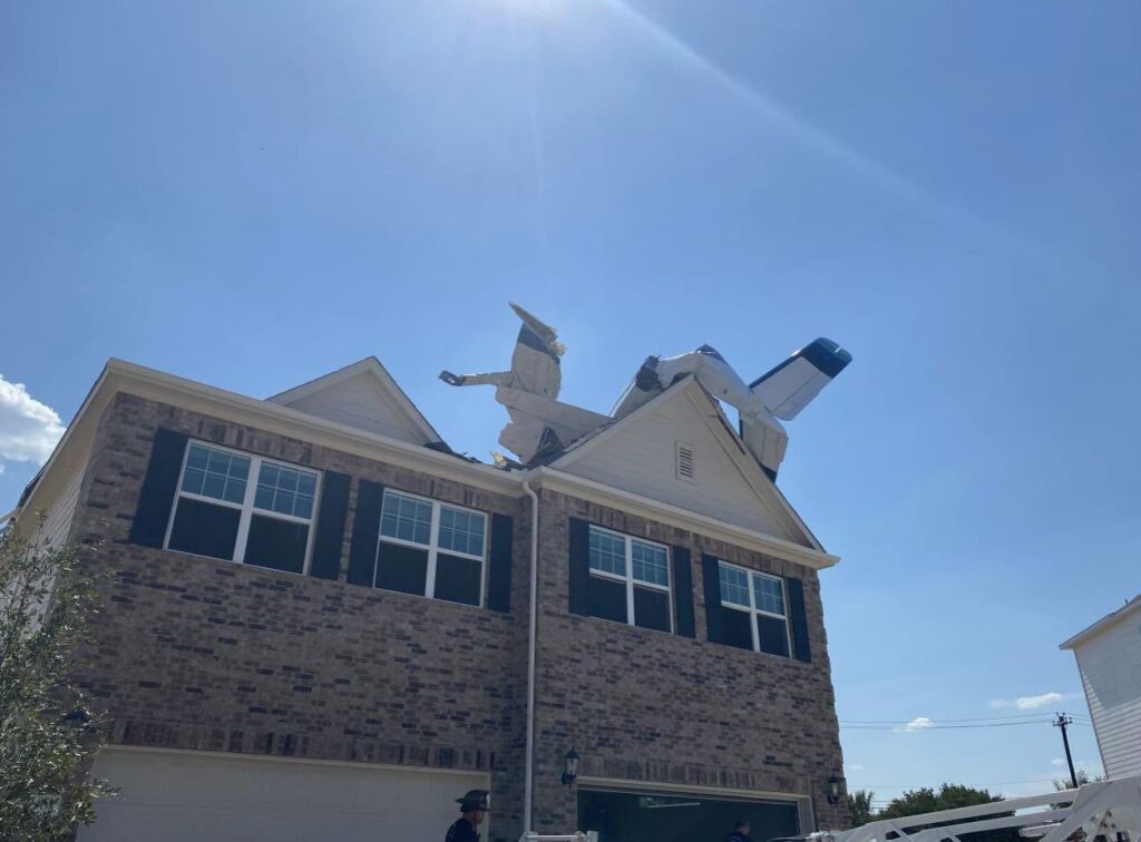 Wide view showing a light aircraft crashed into the roof of a two-story brick house, the tail section sticking upward into the sky while the nose is buried inside the roof structure.