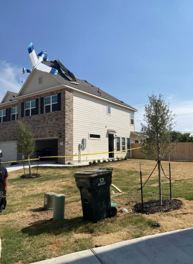Street-level view of a suburban home with a small aircraft lodged in the roof; yellow caution tape surrounds the scene as responders and bystanders observe from the driveway.