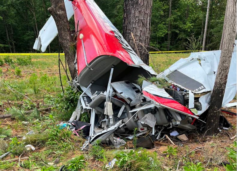 A close-up view of the crashed airplane shows the crushed fuselage and cockpit wedged against two trees. The wings and structure are severely deformed, with debris and vegetation mixed around the impact site.