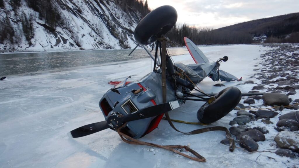 A gray and red Piper PA-18 Super Cub lies inverted on a frozen riverbank with its landing gear up in the air. The aircraft’s propeller is bent and partially buried in ice, and the fuselage shows significant deformation. Snow and rocks line the river’s edge, with steep, forested cliffs visible in the background under an overcast sky.
