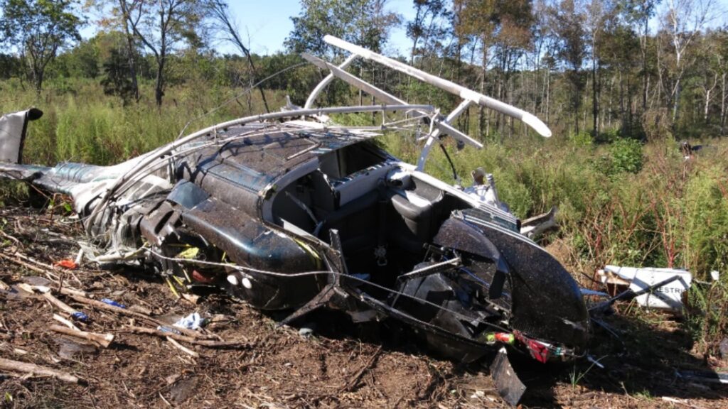 Close-up of the helicopter fuselage lying on its right side in a brushy field, showing severe structural damage, missing windscreen sections, and broken rotor components surrounded by scattered debris.