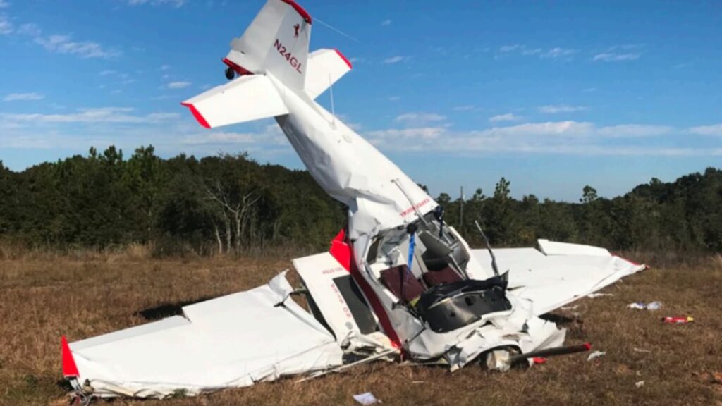A heavily damaged white Bushby Mustang II aircraft, tail number N24GL, sits nose-down in a grassy field. The fuselage is crushed, the tail is vertical, and both wings are bent and torn. Bright blue sky and scattered clouds appear overhead with trees in the background.