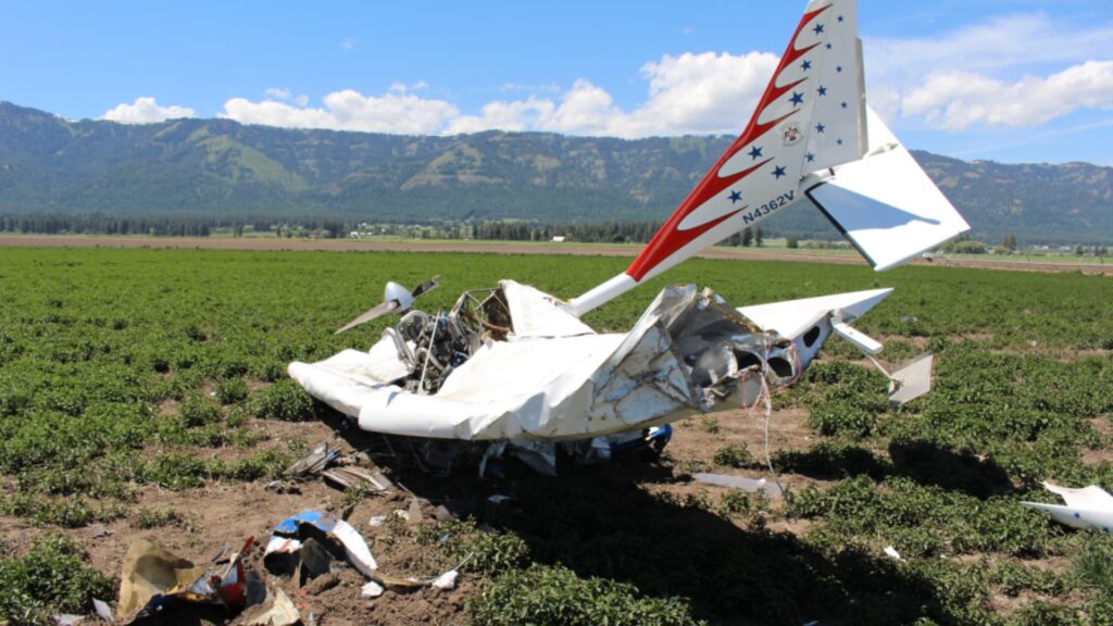 Wreckage of an experimental Titan II aircraft lying in a farm field, showing a severely crushed fuselage, collapsed wings, and exposed engine components. The tail section with registration number N4362V remains mostly intact and upright. Mountains and open farmland are visible in the background under a clear sky.