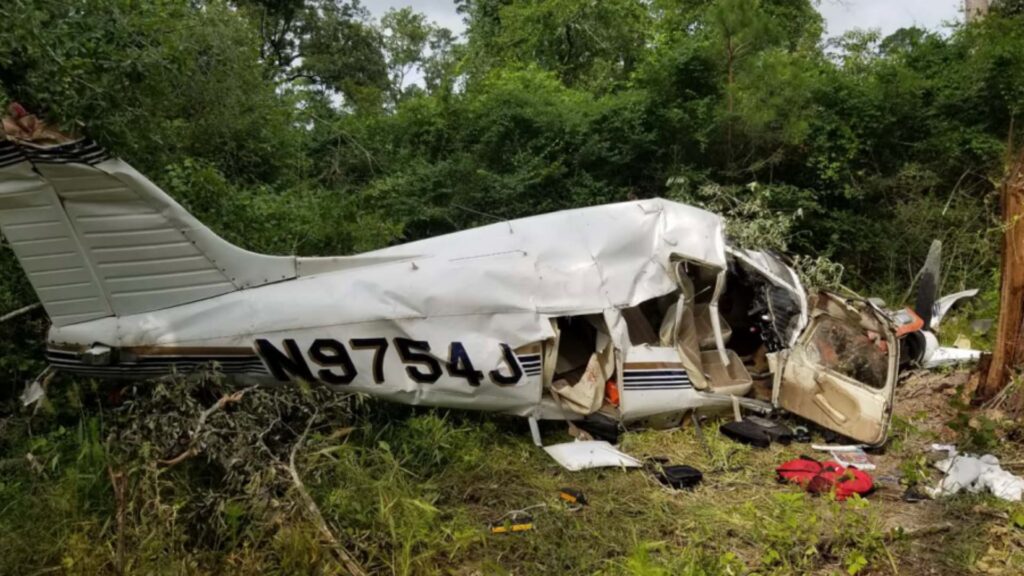 A heavily damaged Piper PA-28-180 aircraft rests on its right side in a wooded area. The fuselage is crushed and buckled, the cabin structure is destroyed, and debris is scattered on the ground. The registration number N9754J is visible along the side. The surrounding vegetation is dense, with broken branches and disturbed brush around the wreckage.