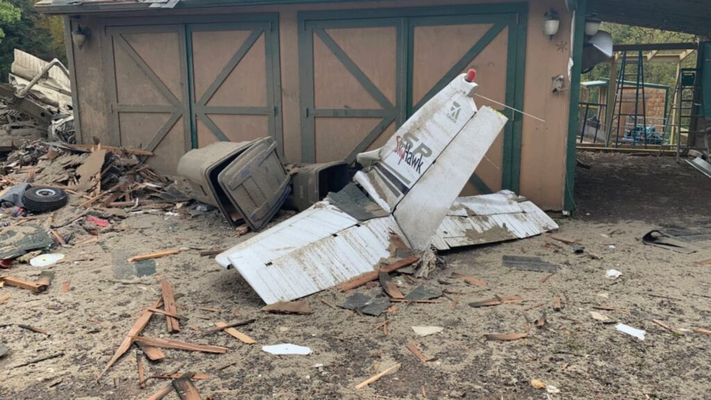 The tail section of a Cessna Skyhawk airplane lies against a damaged garage with green-trimmed wooden doors. Wreckage and scattered debris cover the ground nearby.