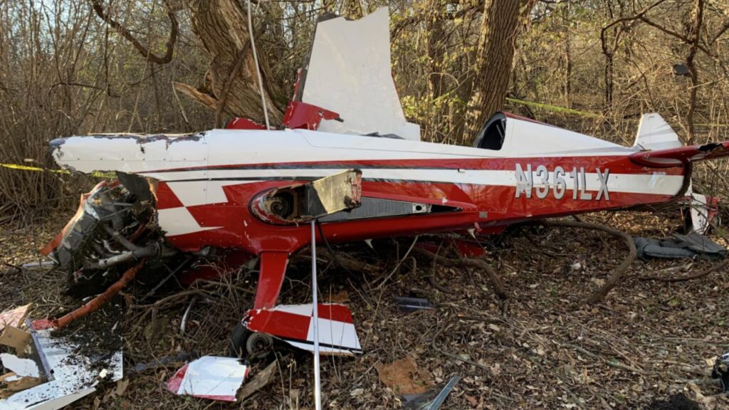 Side view of the crashed Extra EA300/LC showing registration number N361LX; the fuselage is mostly intact but the engine compartment and wings are heavily damaged among trees.