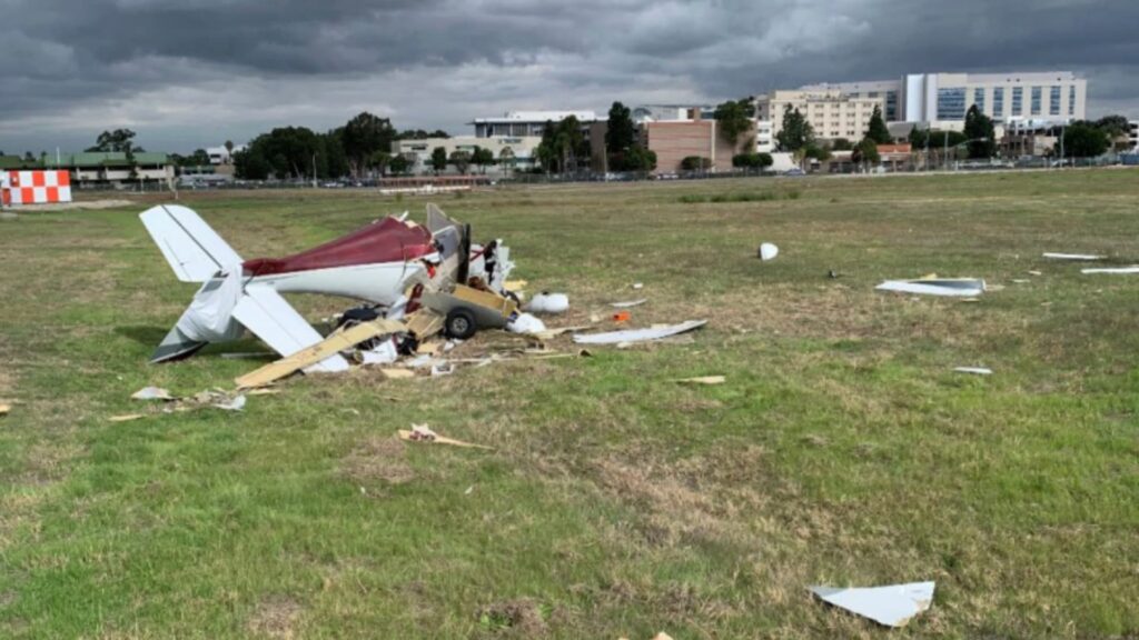 Wreckage of a small airplane lying in a grassy field near an airport. The aircraft is severely damaged with the fuselage broken apart and debris scattered around. The tail section remains partially intact, and dark storm clouds hang overhead with buildings visible in the background.
