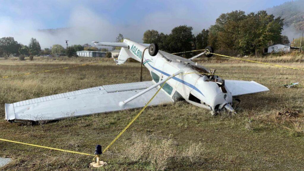 A white Cessna 172 airplane with registration N1870V is shown inverted on grassy terrain near the edge of a runway. The nose is crushed, the propeller bent, and the wings and tail are mostly intact but dirt-covered. Yellow caution tape surrounds the wreckage, and light morning fog and trees are visible in the background near a small building.