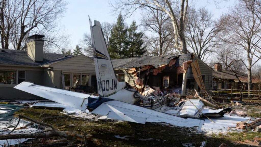 Wreckage of a Piper PA-31-350 aircraft embedded in the side of a residential house in Madeira, Ohio. The tail section, marked N400JM, remains mostly intact while the fuselage and left wing are heavily damaged. The roof and wall of the home show severe impact damage, with debris scattered across the yard.