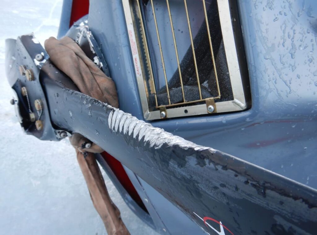 Close-up view of the damaged propeller on the crashed Piper PA-18. The blade shows deep, uniform gouges consistent with a wire strike, and the surrounding cowling and engine area have visible impact damage. The metal surface is wet and icy.