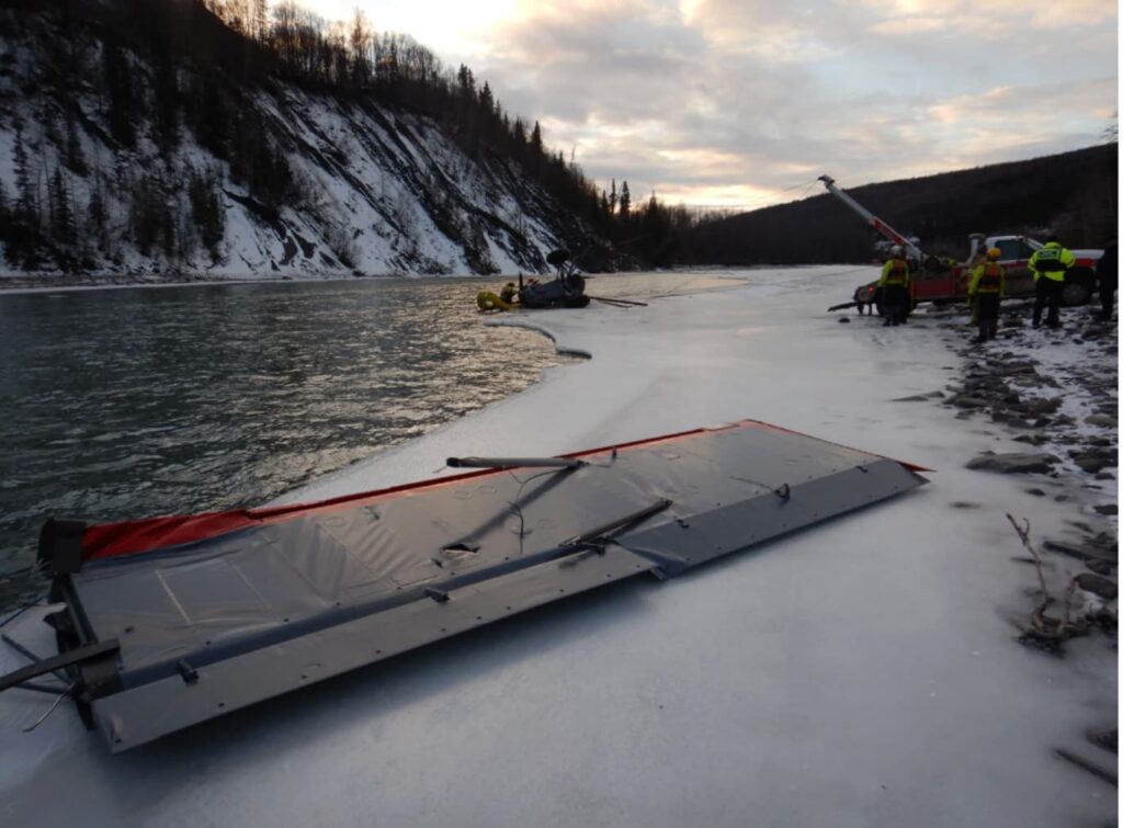 A separated wing from the Piper PA-18 lies on a frozen river near first responders and a recovery truck working along the shore. In the distance, the inverted fuselage of the aircraft remains partially on the ice and in the water. Snow-covered cliffs and trees line the river beneath a cold, cloudy sunset.
