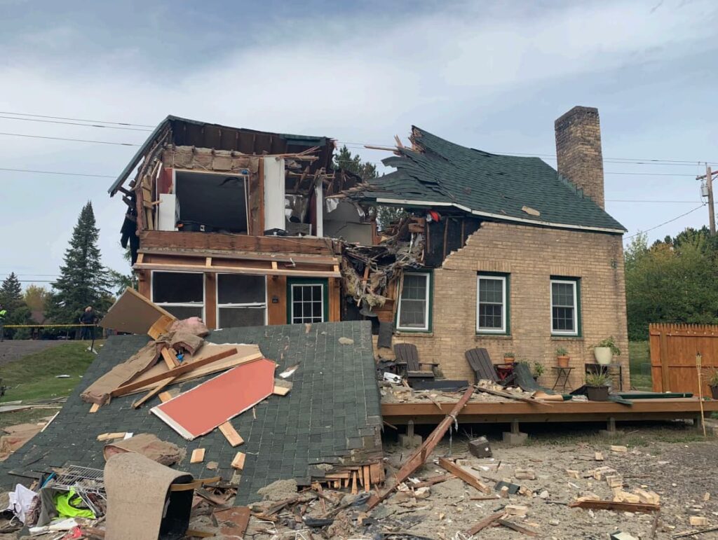 A two-story brick house severely damaged by an aircraft impact. The upper story and part of the roof are destroyed, exposing interior rooms, insulation, and debris scattered in front of the house.