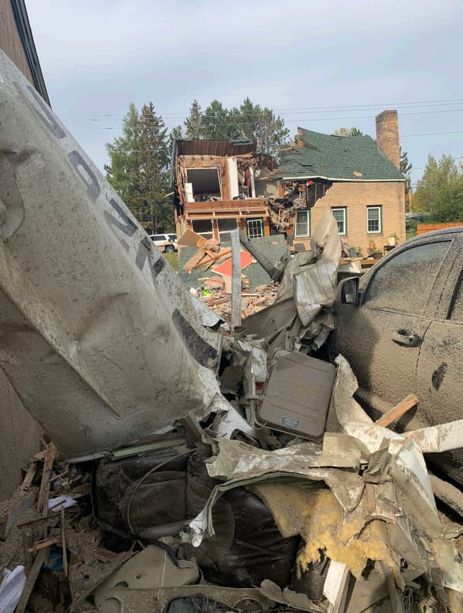 Close-up of crumpled aircraft wreckage and debris pressed between a damaged garage and a vehicle, with the destroyed house visible in the background.