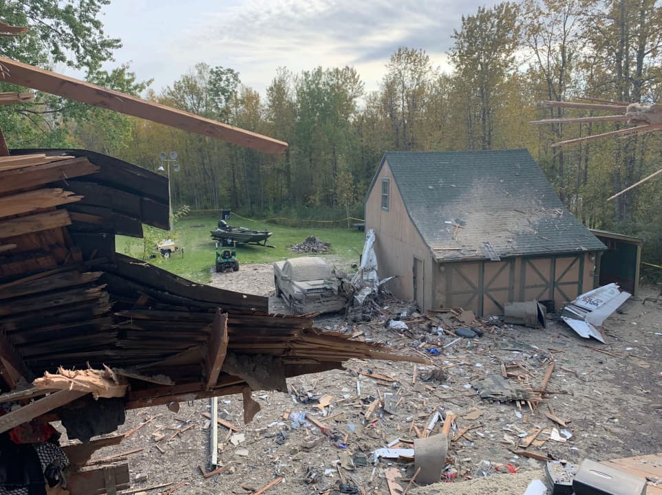 View from the upper story of a destroyed house showing scattered debris, a heavily damaged garage, and wooded trees beyond the crash site.