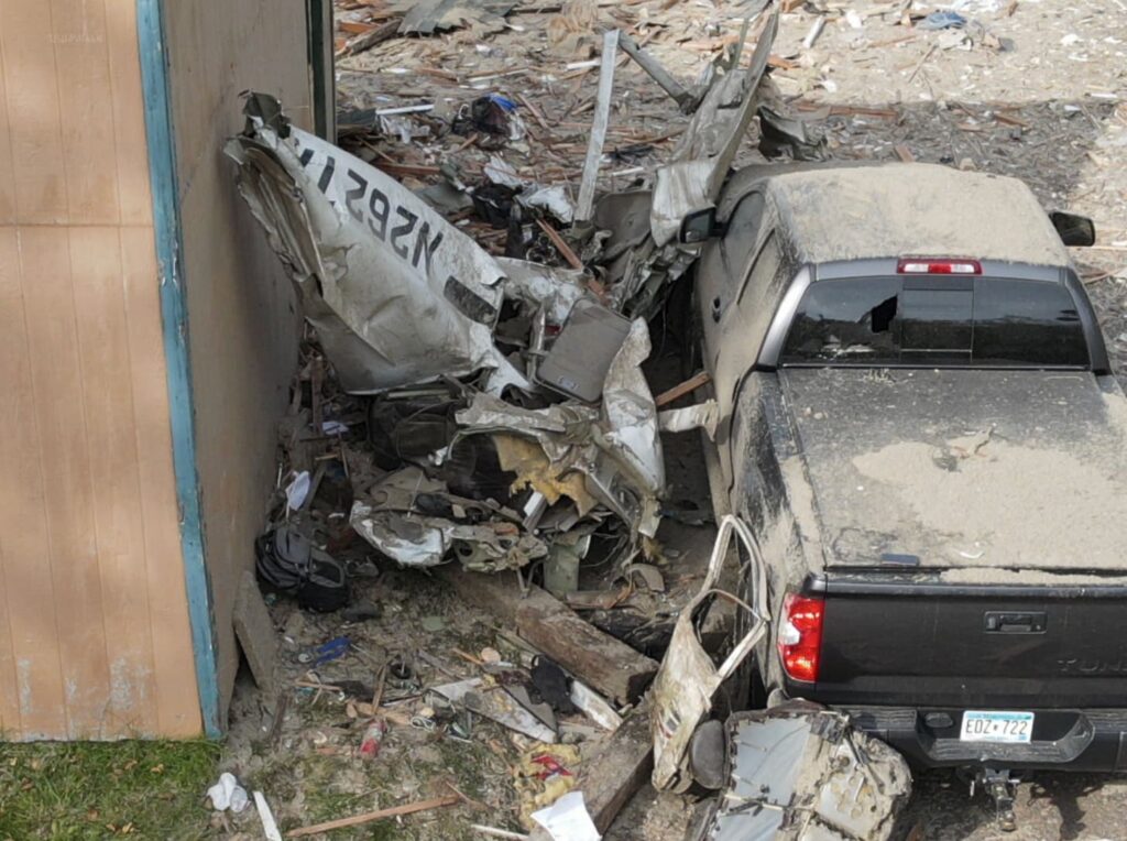 Aerial view of a black pickup truck and the crushed fuselage of a Cessna aircraft marked N262TA wedged between the truck and a garage wall, surrounded by wreckage.